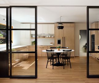 A kitchen with wood and white cabinets, white countertops, and a wooden floor. The kitchen is open plan with an attached dining room and living area, but glass screens and doors give the option to divide the space