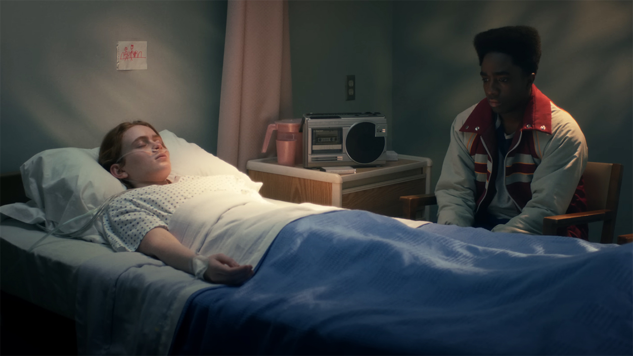 A comatose Max lying in a hospital bed as Lucas sits next to her in Stranger Things season 5