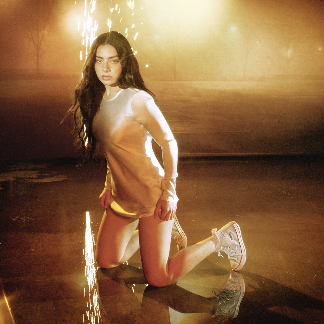 Charli kneels on the floor while wearing a long-sleeve metallic dress and Chuck Taylor All Star shoes