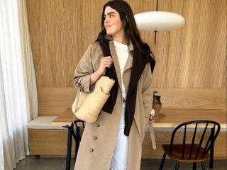 British fashion influencer Anna Howard poses in a modern London kitchen wearing a trench coat, black sweater draped over her shoulders, white t-shirt, belt, white pants and straw tote bag