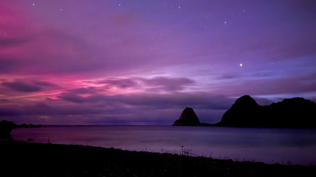 image of a beach at night with distant mountains and a partly cloudy sky illuminated with pinks and reds.