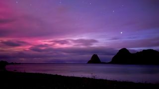 image of a beach at night with distant mountains and a partly cloudy sky illuminated with pinks and reds.