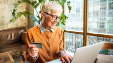 A smiling older woman holds a bank card in her hand while typing on a laptop