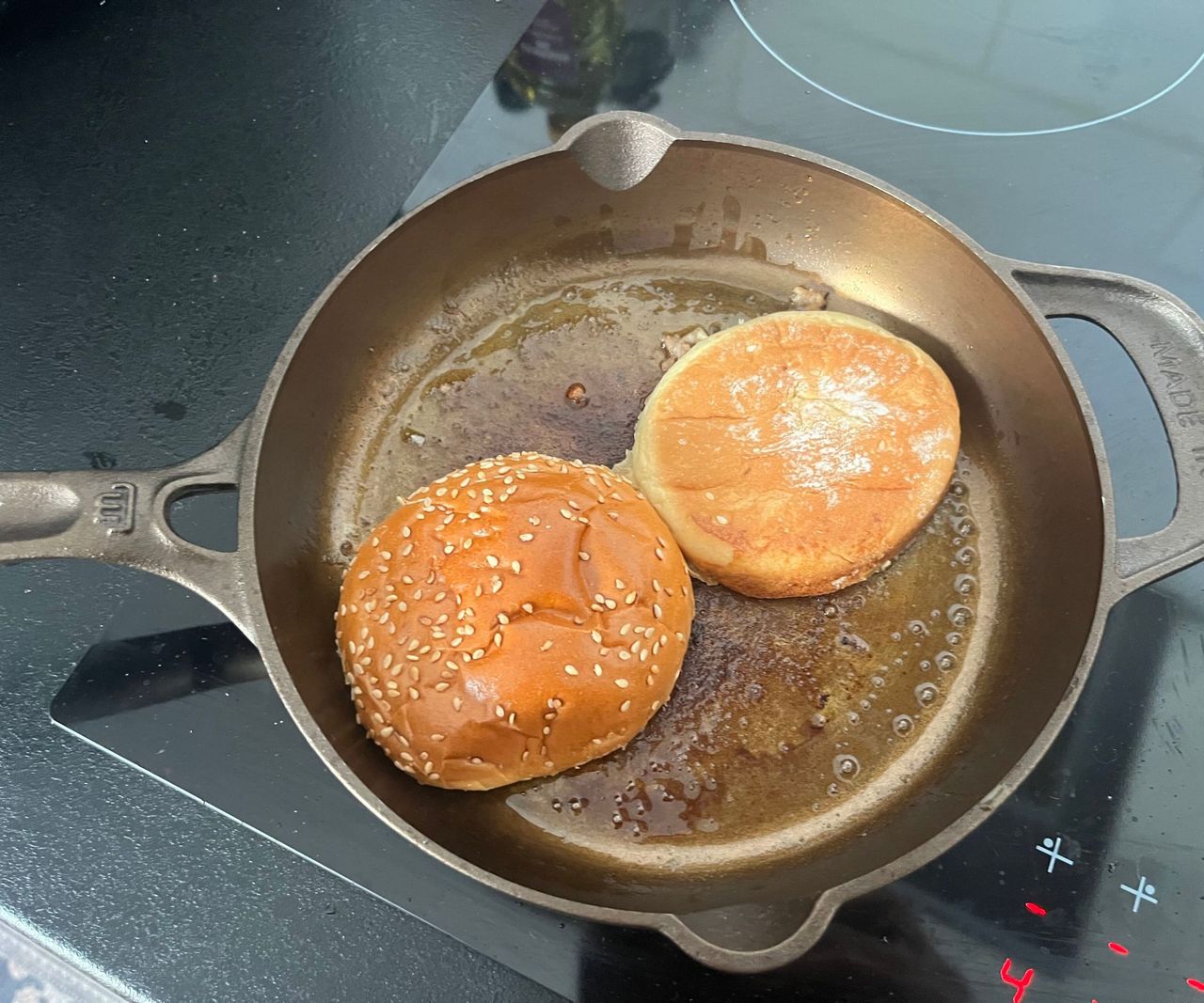 A small bronze colored cast-iron skillet on a black glass stove with a burger bun toasting inside