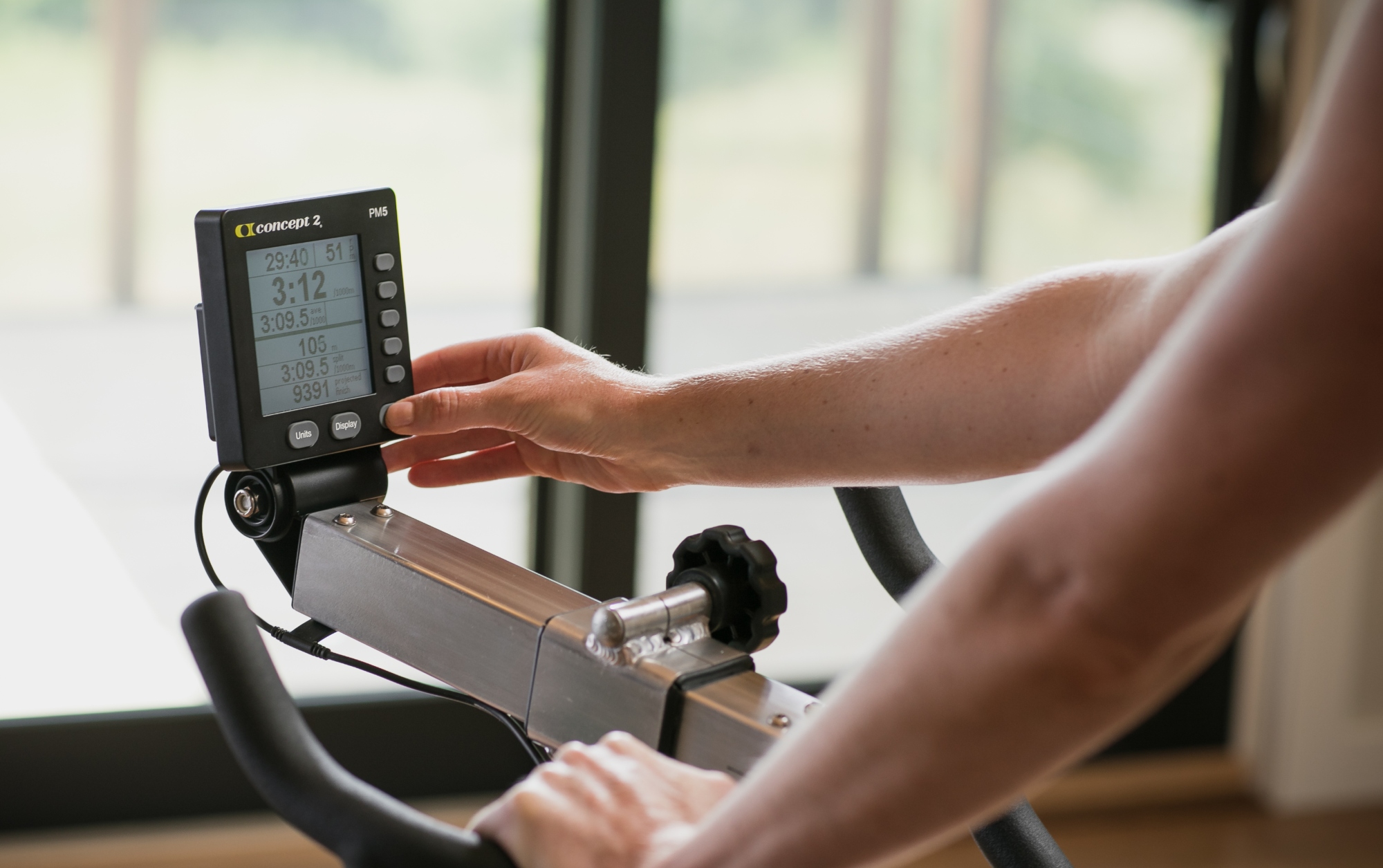 This image shows the handbars of a bike and a Concept2 BikeErg PM5 monitor being programmed by someones hands