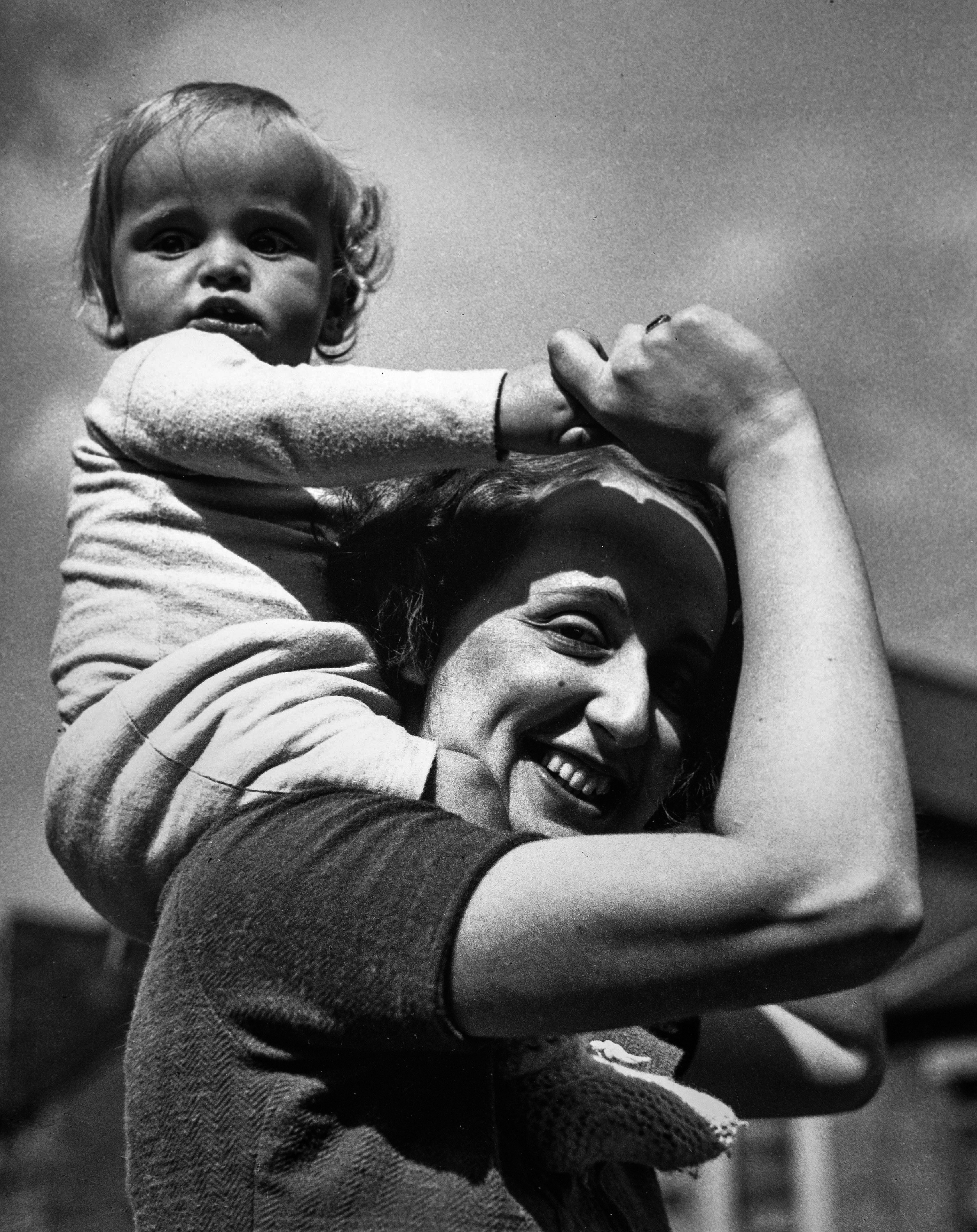 A smiling woman holds a fair-haired toddler aloft on her shoulders in bright sunlight, gripping the child's small hands as they reach upward. The woman looks up with an open, joyful expression. The image is warm and spontaneous in feeling.