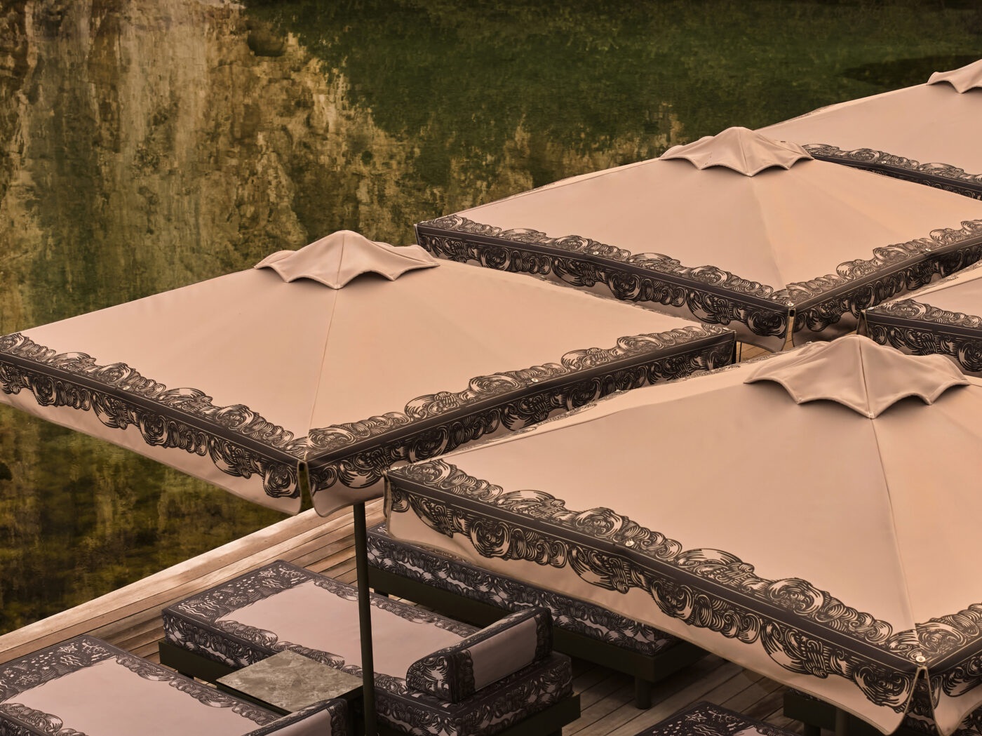 A series of sun loungers carved from olive green-painted wrought iron and pale rose textile decorated with blue hand-drawn natural motifs sits on a wooden pavement in a sun-lit beach club.