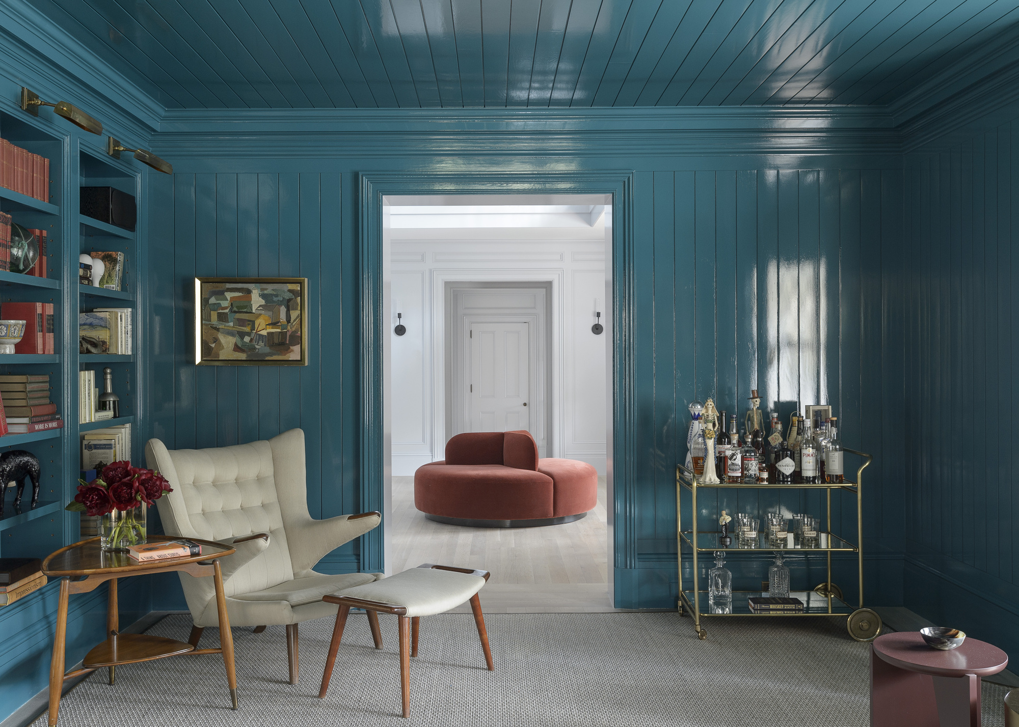 A home library with panelled walls color drenched in teal in a gloss finish, a built-in bookshelf, and a bar cart in the corner