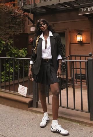 A woman wearing a black pleated miniskirt with a white button-down shirt, a black blazer, and sneakers.