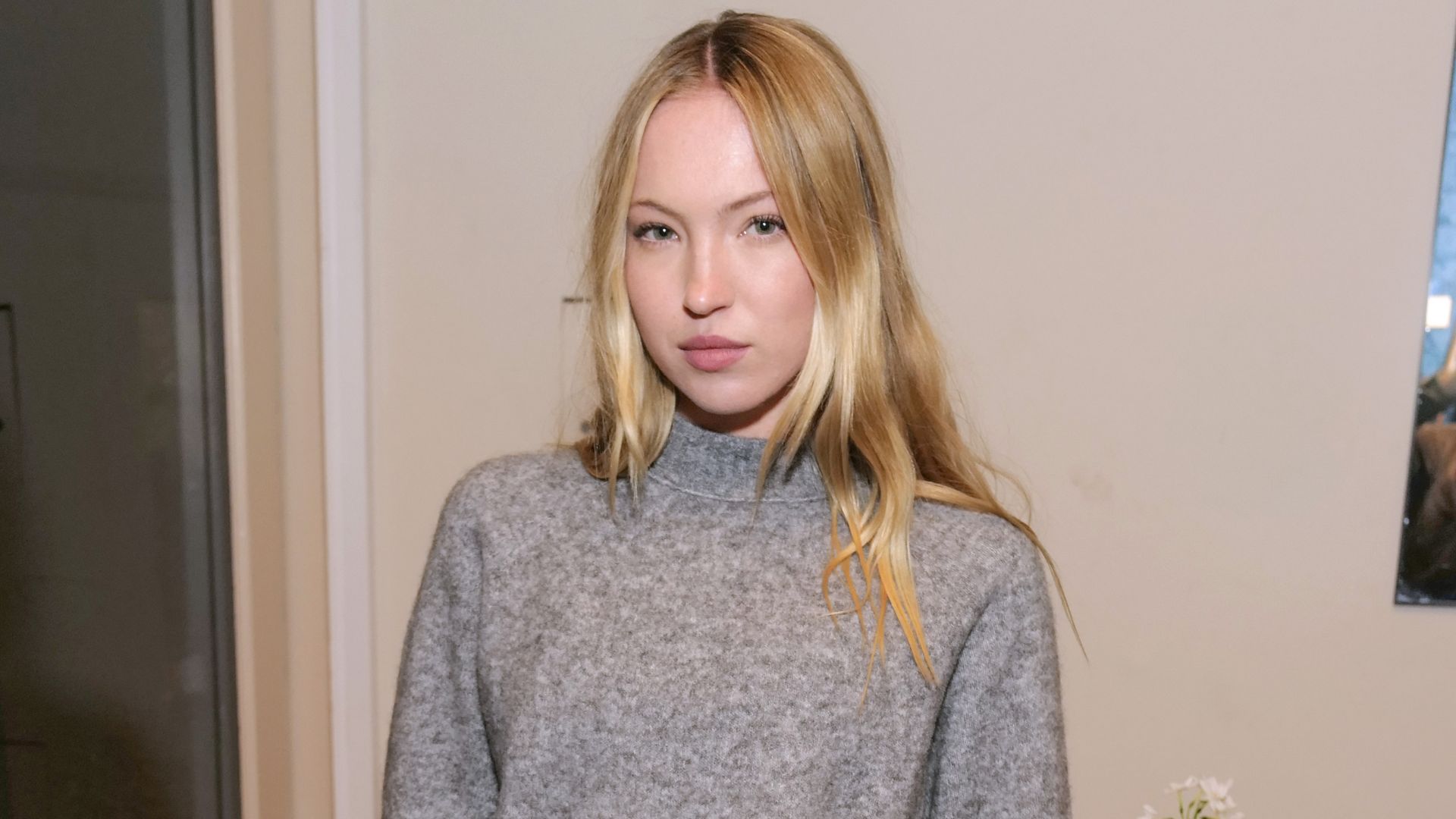 Lila Moss poses for a photo wearing black capri pants with a grey jumper, an oversized belt and black slingback heels.