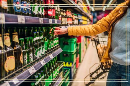 A close up of a person reaching for a bottle of beer on a supermarket shelf