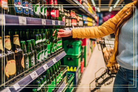 A close up of a person reaching for a bottle of beer on a supermarket shelf