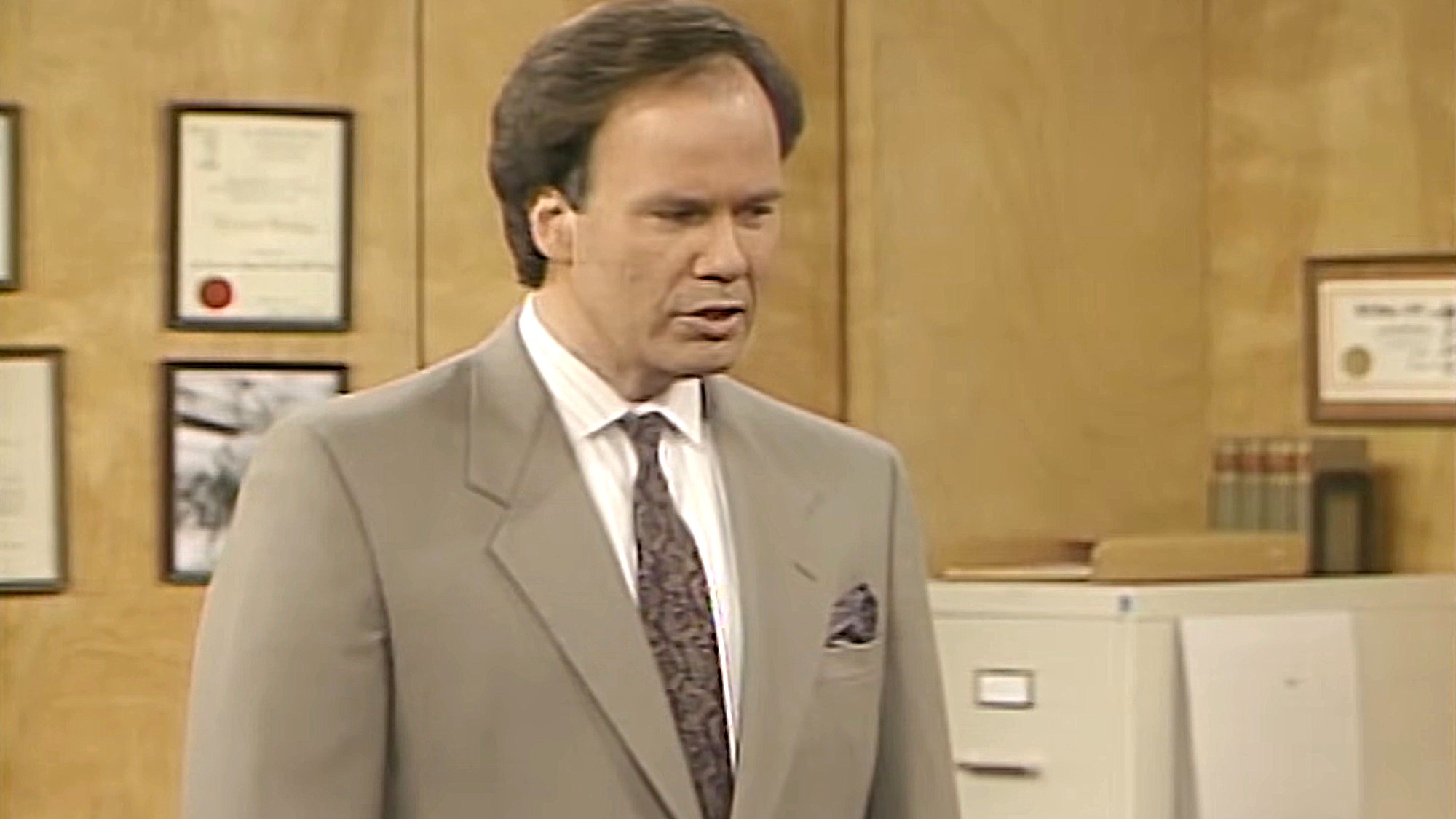 Dennis Haskins is shown as Mr. Belding on Saved by the Bell, wearing a brown suit as he stands in his office. A filing cabinet and framed certificates are shown in the background.