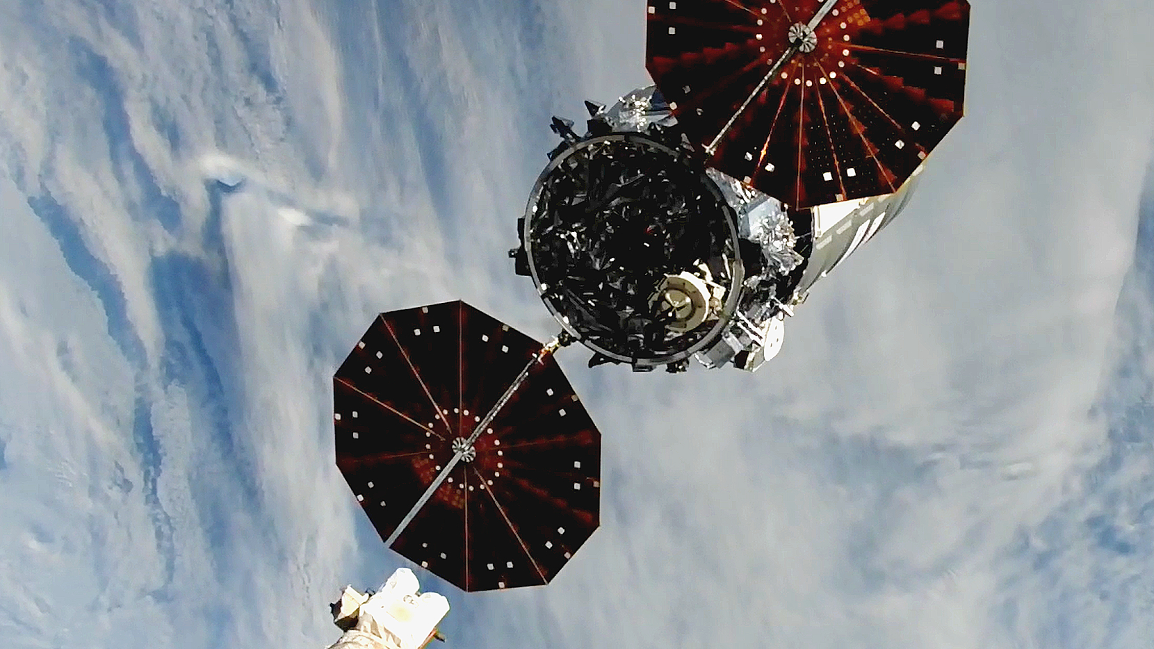 In the foreground is a large cargo ship carrying two large solar panels on its sides, and the Earth in the background with clouds and blue peaks of the South Atlantic Ocean.