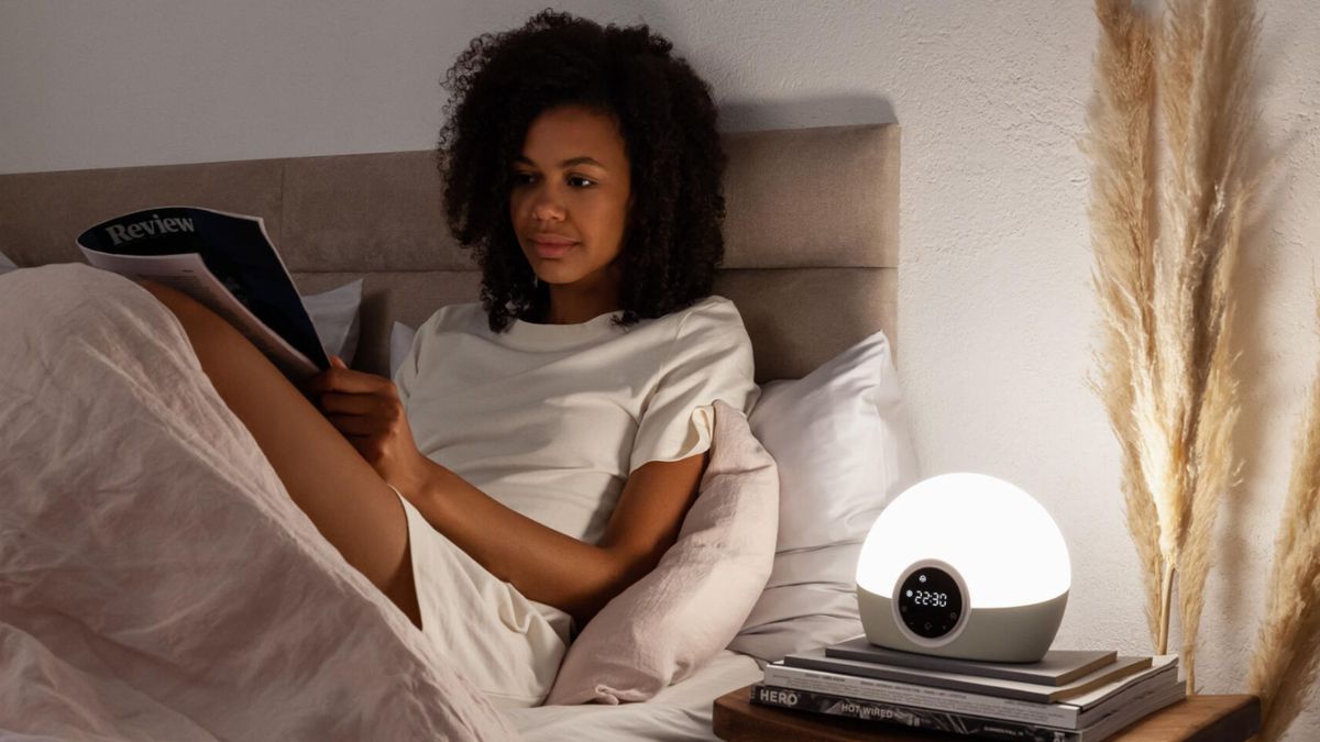 A woman sitting in bed reading next to a Lumie sunrise alarm clock on top of stack of books on bedside table and pampus grass as she becomes a morning person