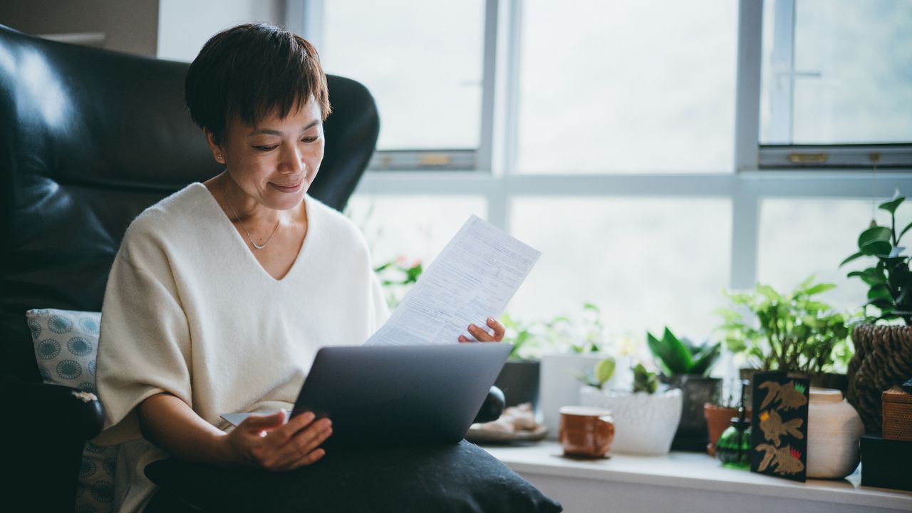 An older woman looks at paperwork with her laptop on her lap while she sits in a big chair.