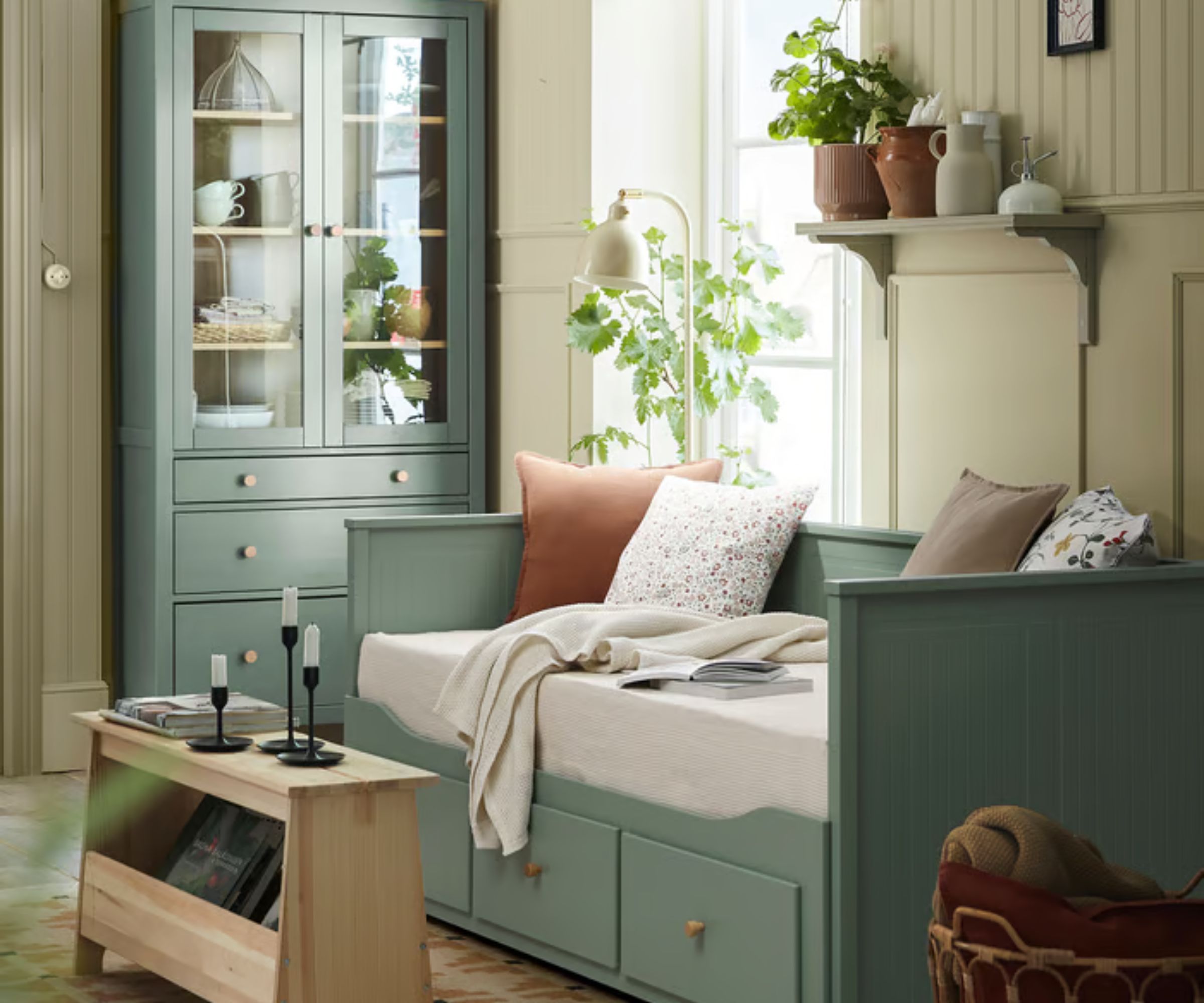 IKEA sage green HEMNES bed with neutral cushions, a wooden side table with black candlesticks, a sage green painted dresser with ceramics and tableware and a wood panelled wall painted cream.