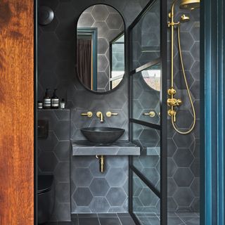 A dark bathroom scheme with black hexagonal tiles, oblong mirror, black matt basin, and-brass showerhead