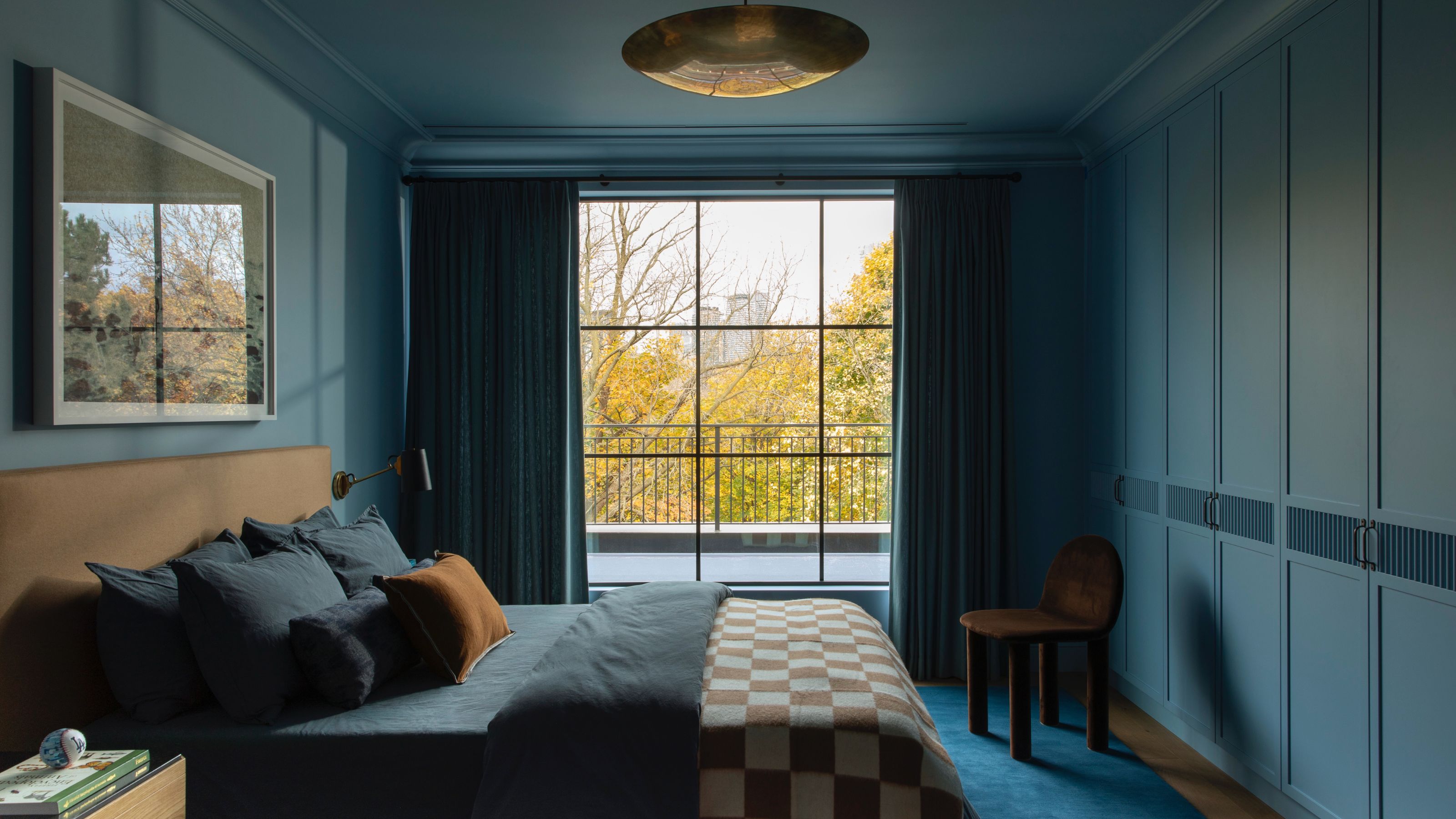 A moody and modern bedroom fully saturated in deep blue tones on the walls and built-in wardrobes. It features a large black-framed window overlooking autumn trees, a bed with a tan headboard and navy bedding, and a brown velvet accent chair