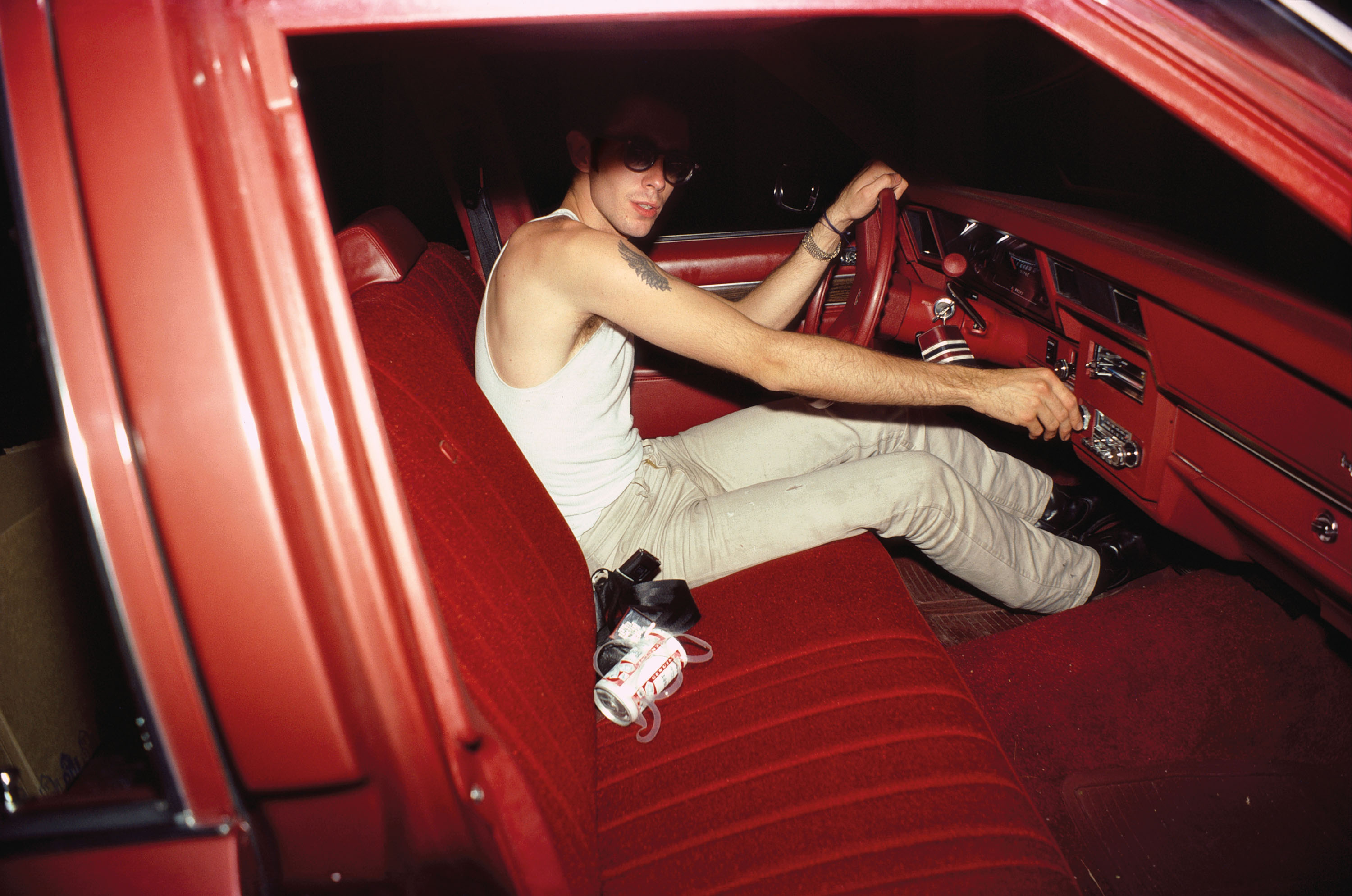 A man wearing sunglasses and a white tank top sits in the driver's seat of a car with a vibrant red interior, reaching toward the dashboard.A man wearing sunglasses and a white tank top sits in the driver's seat of a car with a vibrant red interior, reaching toward the dashboard.