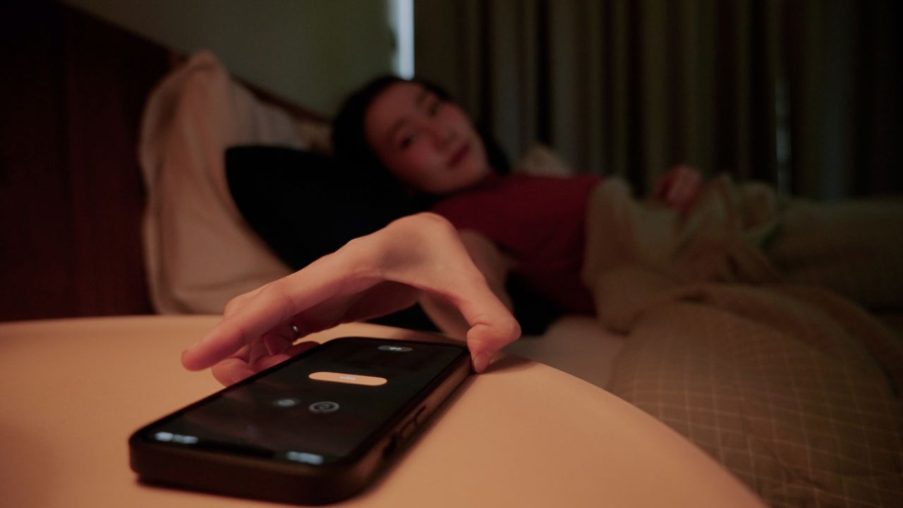 A woman in bed reaches for a smartphone on a nightstand. The phone screen shows an alarm going off.