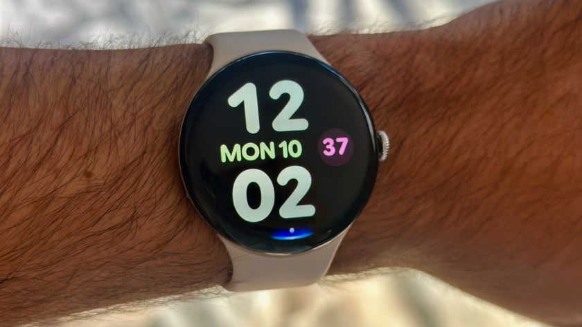 A photo of the Google Pixel Watch 4 on a man&#039;s wrist showing the Bold Utility watch face with large, colorful clock numbers and day/date info. At the display bottom is a blue Gemini Raise to Talk indicator.