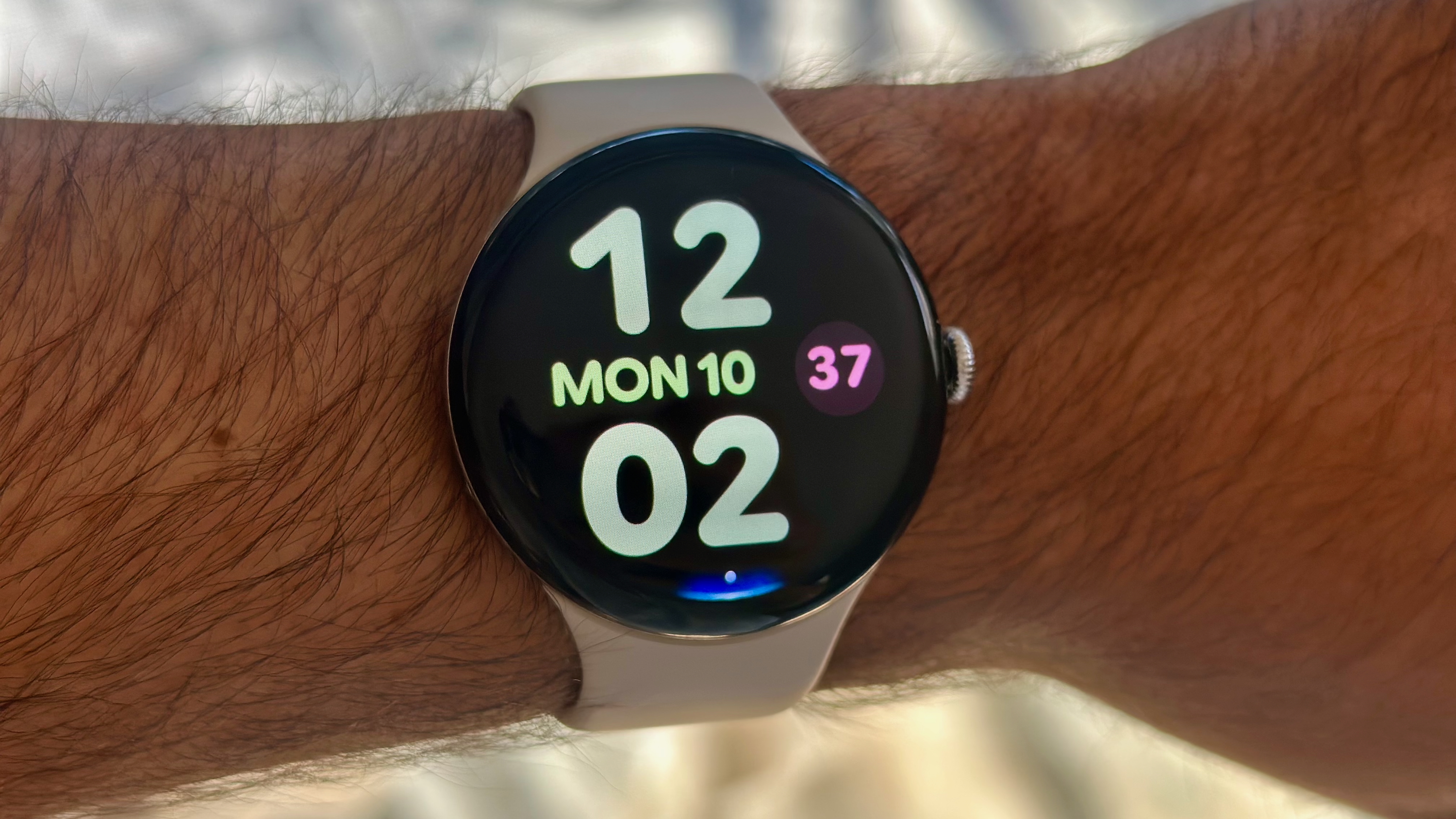 A photo of the Google Pixel Watch 4 on a man's wrist showing the Bold Utility watch face with large, colorful clock numbers and day/date info. At the display bottom is a blue Gemini Raise to Talk indicator.