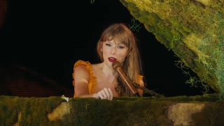A screenshot of Taylor Swift talking into a mic while sitting behind a piano during the trailer for The Eras Tour concert film.