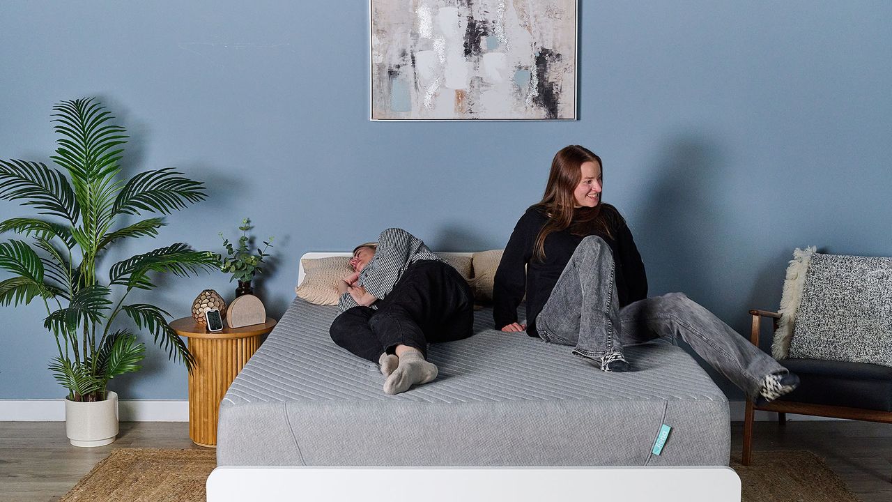 This image shows our subjective motion isolation test, conducted in our sleep studio. In the middle of a decorated room is the Siena Mattress on a bed frame. On the left of the mattress, sleep editor Nicola Appleton rests in a side sleeping position. On the right, sleep writer Frances Daniels sits up on the mattress displaying a moderate force movement.
