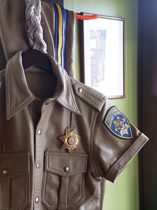 Close up of a California highway police patrol leather beige colour short sleeve shirt, police badge, hanging from a rope attached to a red rail, with matching leather pants draped behind, framed picture on a lime green and brown wall in shot