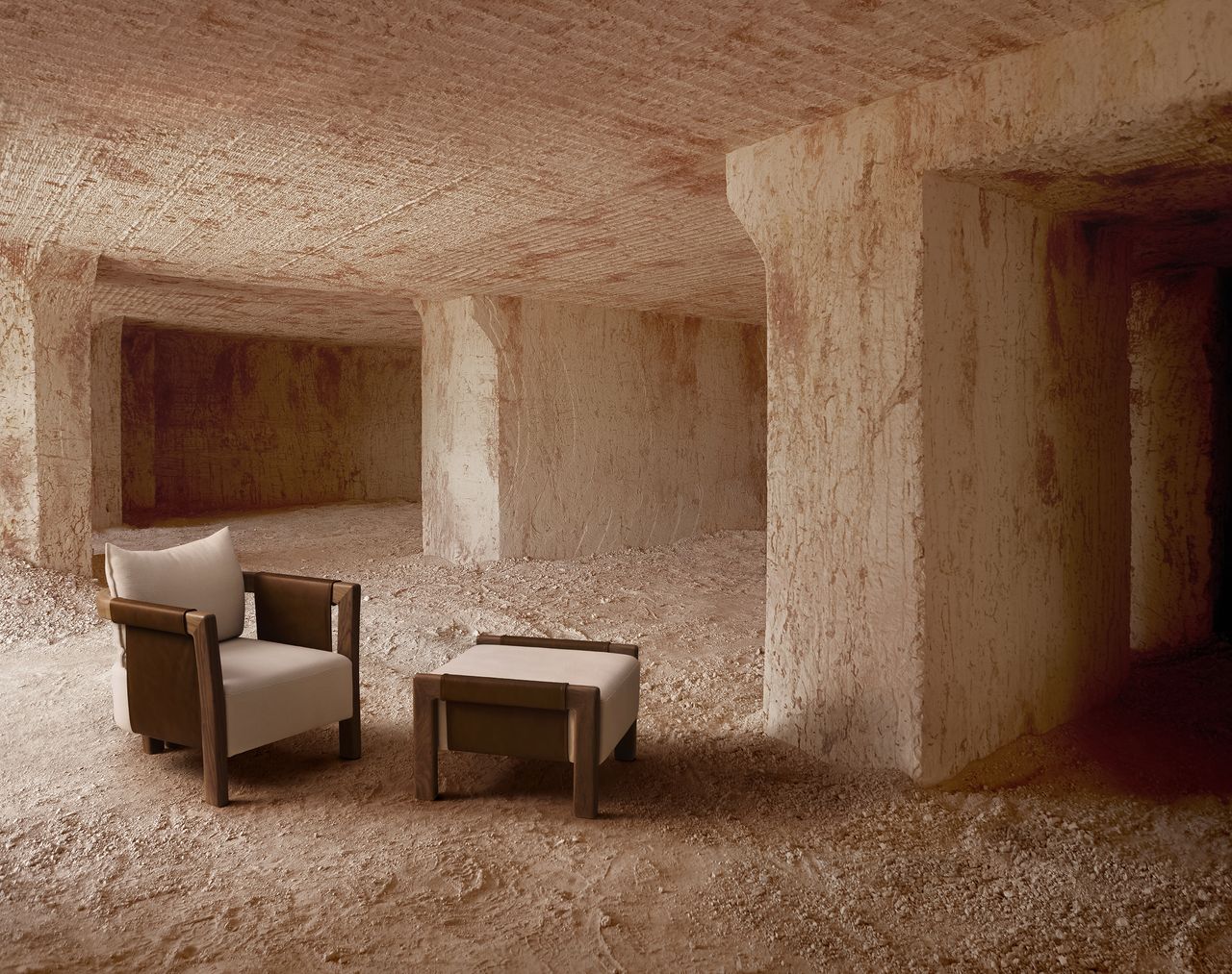 In an exposed cement, earthy building filled with sand, a mid-century modern-style armchair in wood and leather and a matching Ottoman sit in a bare environment.