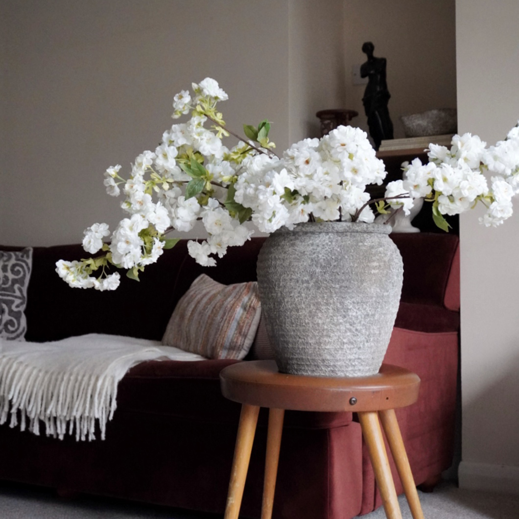 Large grey vase of white flowers on wooden stool beside burgundy sofa