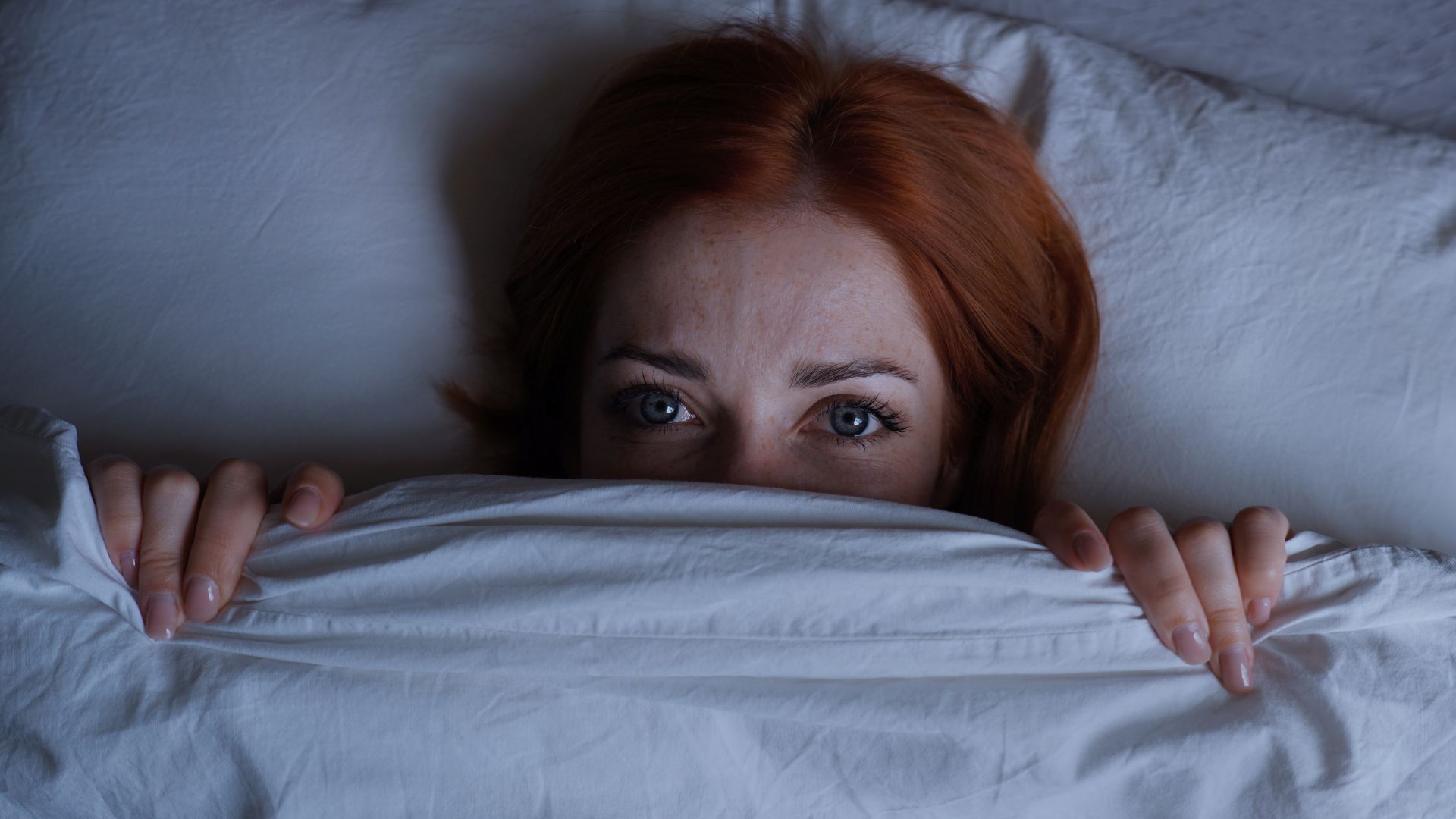 A woman lying on her back in bed, awake at night with the cover pulled up over her chin.