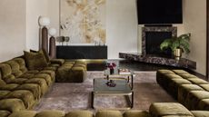 Image of a luxury yet cozy living room. The walls are a white limewash, there is a large olive green sectional that wraps around the room, and the floor is a dark black wood. There is a marble fireplace in the corner with a TV above it.