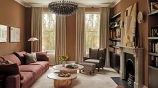 A warm, earth-toned living room featuring terracotta walls, a dusty rose sectional sofa, and a large tiered glass chandelier. Two organic-shaped wooden coffee tables sit on a plush beige rug before a marble fireplace.