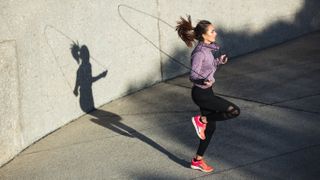 Woman jumping rope with right knee bent while jumping outdoors against a wall backdrop