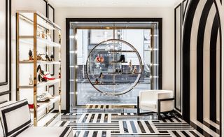 To the left, a gold metal shelf has a lot of shoes displayed. In the center is a huge window, with shoes displayed on a circular metal shelf. The room is decorated in black & white striped tiles and white walls with black stripes.