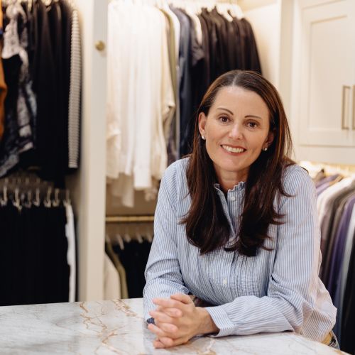 Jenny is a white woman with long dark straight hair, pictured sitting inside a very organized walk-in closet
