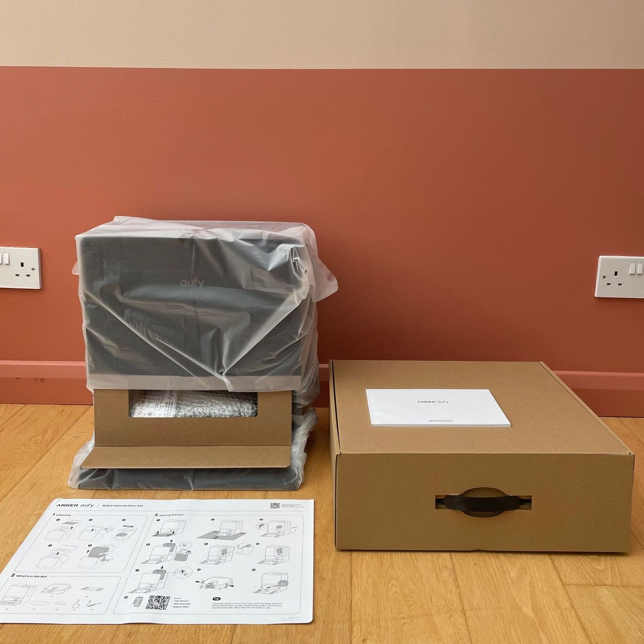 Anker eufy Robot Vacuum E35 Omni components wrapped in plastic and cardboard packaging, on wood flooring in front of a pink painted wall