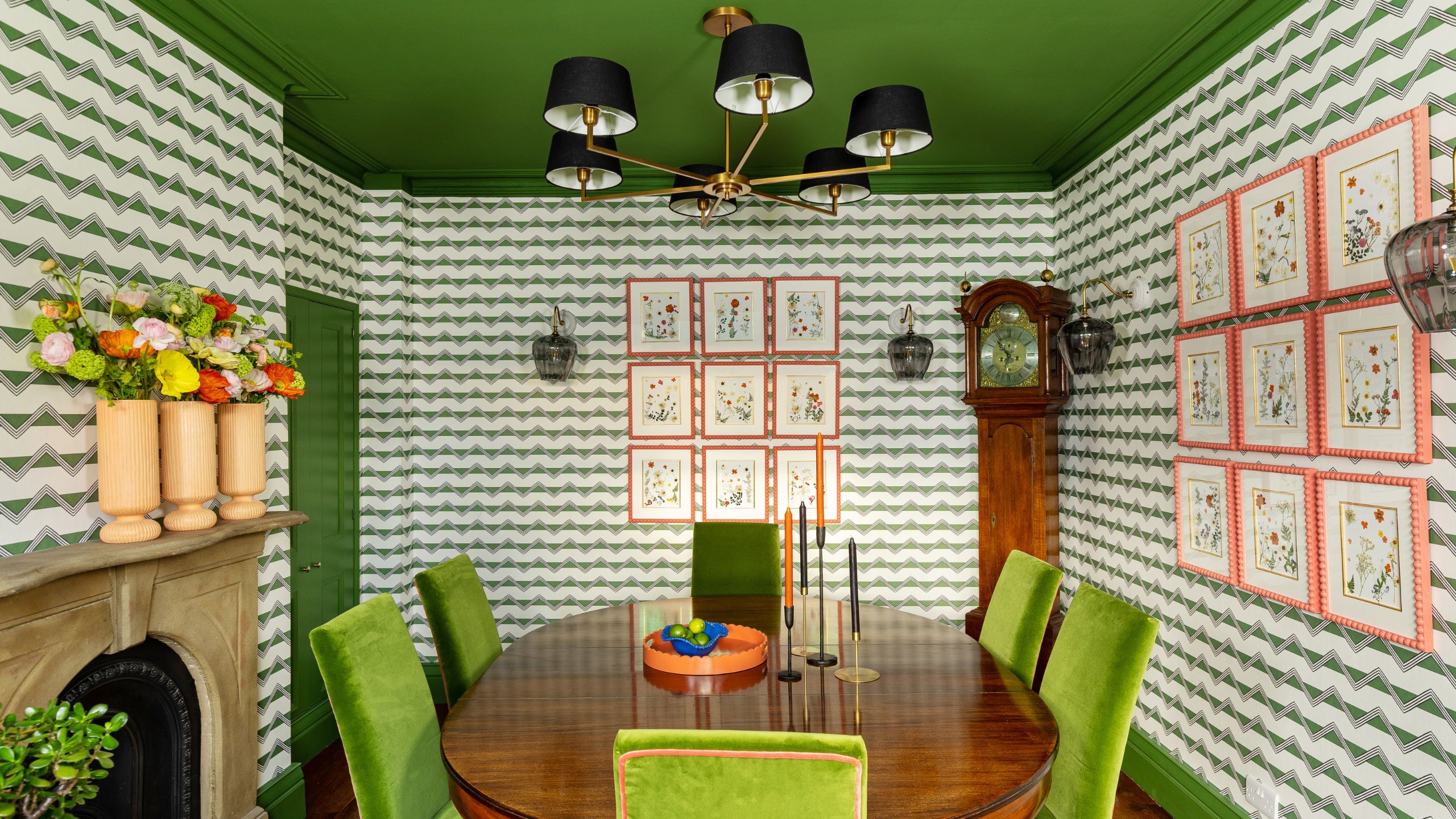 Dining room with green-patterned wallpaper, a painted green ceiling, green velvet chairs around a wooden table, and framed botanical prints.