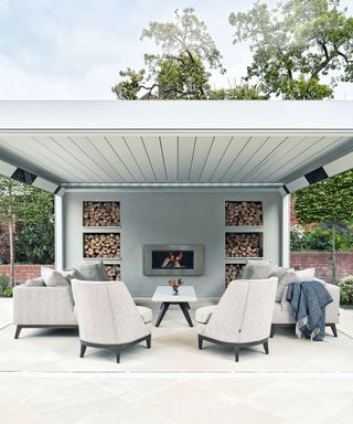 Large outdoor pavilion on a paved terrace, a fireplace niche and log storage, armchairs and sofa, with louvred roof and side panels which can be closed