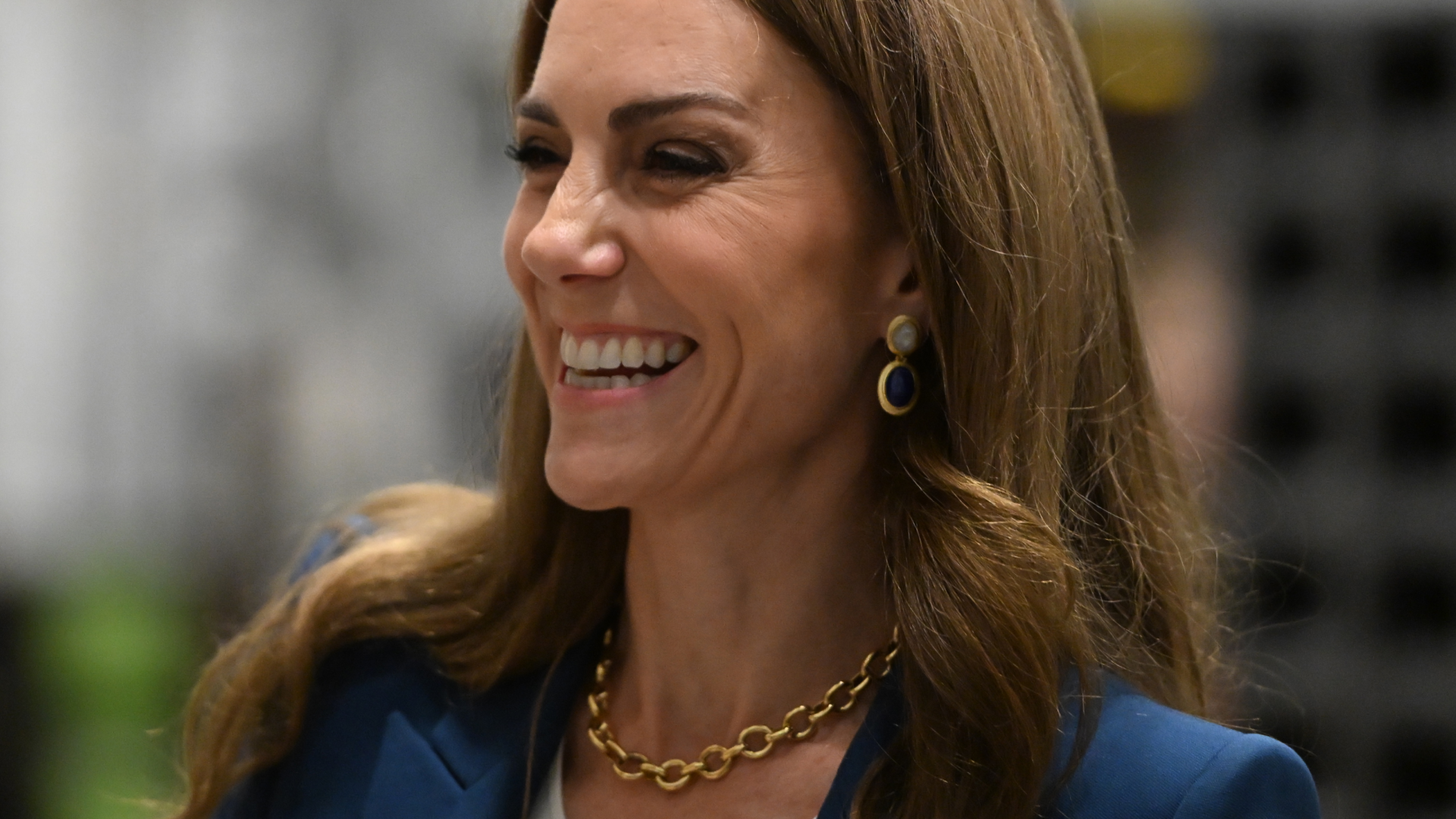 Kate Middleton wearing a blue suit and gold necklace while laughing