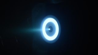 a bright blue-white ring of light on a black background, with a plume of bright white gas ejecting from a nozzle in its center