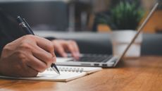 A man makes notes in a small spiral notebook while working on his laptop at his desk, only his hands showing.