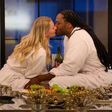 A blond woman (Christine Hamilton) and a black man with long dreads (Vic St. John) kiss while dressed in robes. They sit in floral chairs behind an array of fruit and golden tableware, as the woman holds a green wine bottle between them, in 'Love is Blind' season 10.