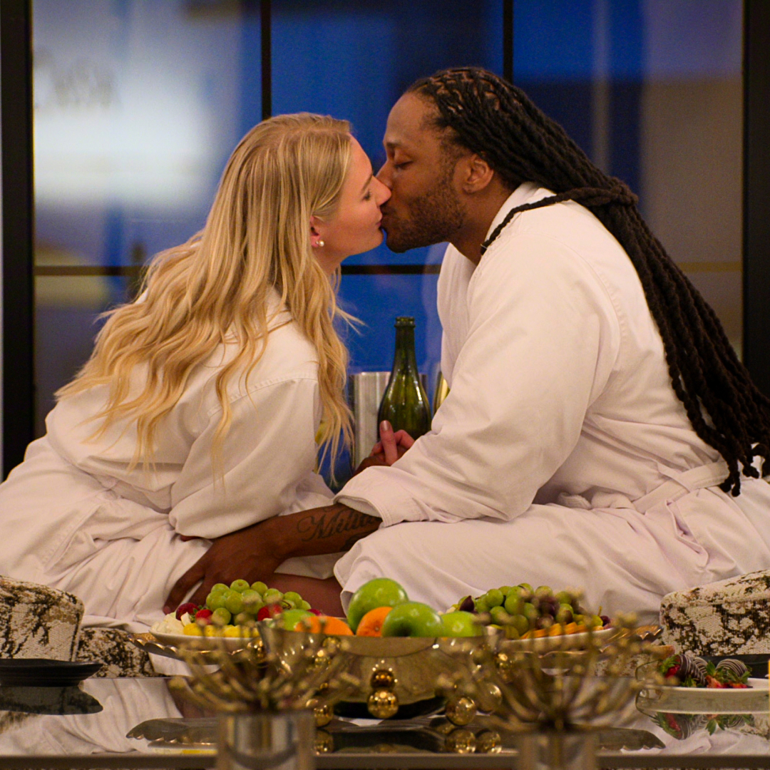 A blond woman (Christine Hamilton) and a black man with long dreads (Vic St. John) kiss while dressed in robes. They sit in floral chairs behind an array of fruit and golden tableware, as the woman holds a green wine bottle between them, in 'Love is Blind' season 10. 