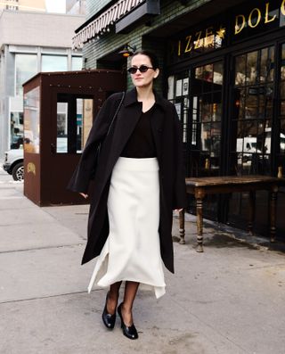 Eliza Huber during NYFW wearing a brown car coat and cashmere sweater, cream skirt, brown tights, and black pumps.