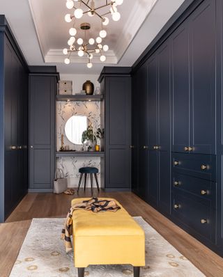blue walk in wardrobe/closet with make up vanity at one end, stool, mirror, wallpaper, yellow ottoman in middle on rug, retro pendant lights