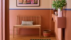 Image of a room painted in a light pink and terracotta pink color block with a royal blue stripe dividing the two paint colors. There is a bench seat, a woven basket, a colorful runner rug in the room