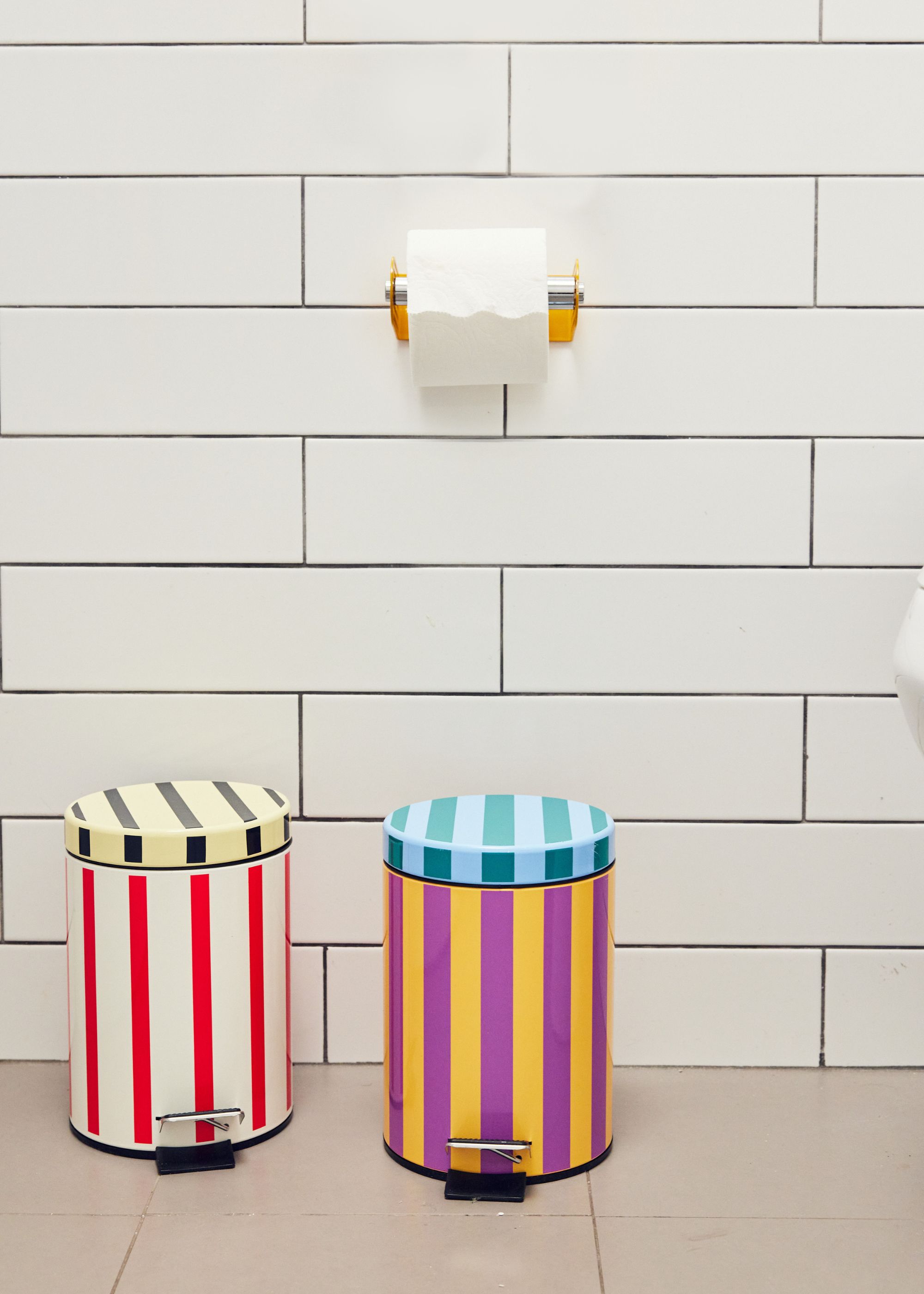 Two colorful and striped trash cans in a bathroom. Above the cans is a toilet roll and in the backdrop is a white tiled bathroom wall.